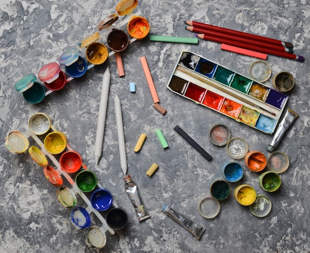 Products for drawing on a concrete table. The concept of inspiration for creation. Multicolored gouache, oil, watercolor paints, crayons, pencils. Top view.