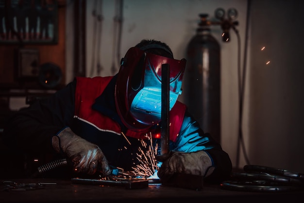 保護ユニフォームを着たプロの溶接工と、工業用ワークショップの後ろにいる他の労働者と一緒に工業用テーブルの金属パイプを溶接するマスク