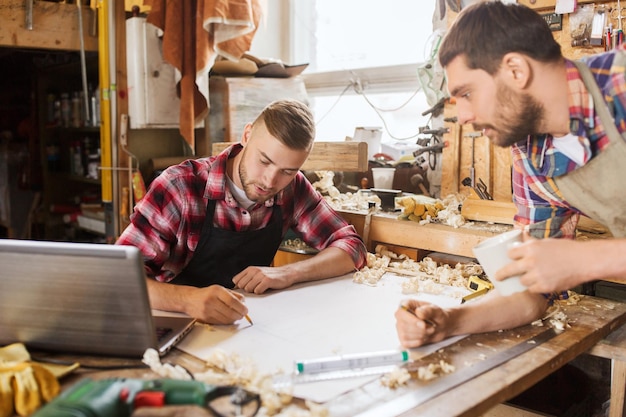 profession, carpentry, woodwork and people concept - two carpenters with laptop computer and blueprint at workshop