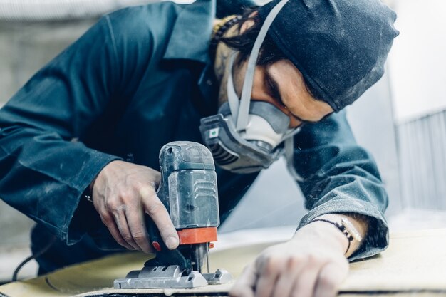 Professione, persone, falegnameria, emozione e concetto di persone: un falegname taglia una tavola con un seghetto alternativo elettrico. fabbricazione di skateboard. ha indumenti protettivi e maschera. 4k