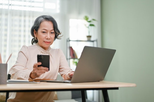 Professional 60s agedasian businesswoman using her smartphone in her office