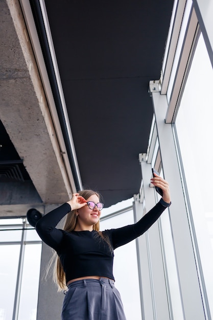 Foto donna d'affari adulta professionista impegnata a mandare sms sul suo telefono cellulare indossando camicia e pantaloni in piedi davanti a grandi finestre di vetro.