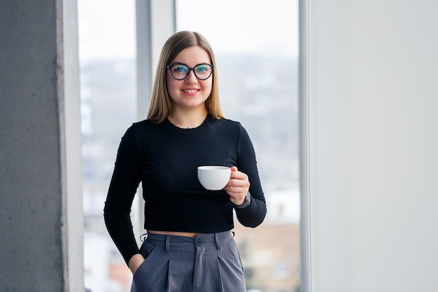 Professional adult businesswoman drinking coffee in shirt and trousers standing in front of large glass windows.