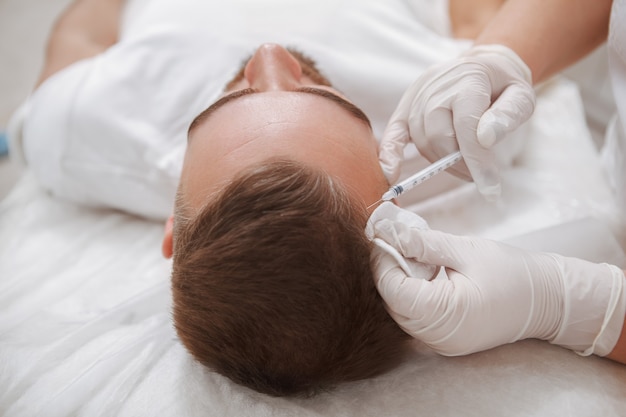 Professional aesthetician giving hairloss treatment to male client, injecting vitamins into his scalp