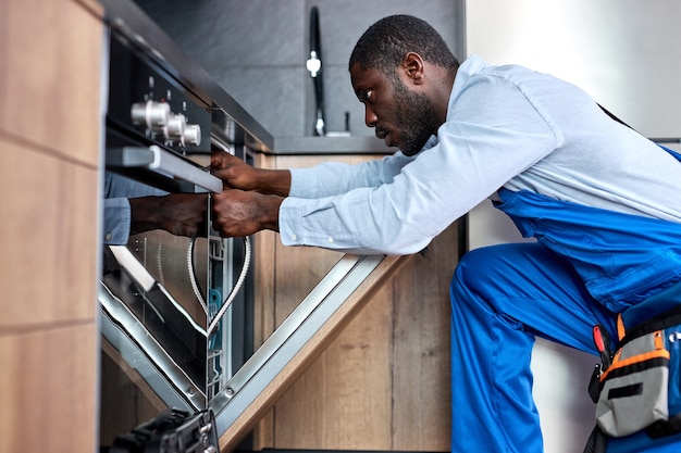 Photo professional afro handyman or contractor repairing dishwasher, need to change old dishwasher hose, black guy in blue overalls is concentrated on work, in kitchen indoors. side view portrait.