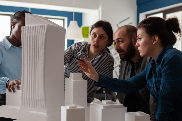 Professional architect with working partners pointing at skyscraper foam scale model in architectural office. Team of engineers collaborating on group project at desk with buildings maquette.