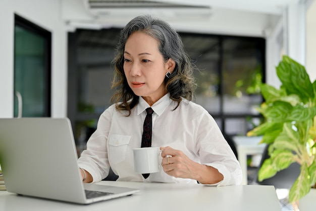 Professional Asian aged female CEO looking at laptop screen and sipping morning coffee