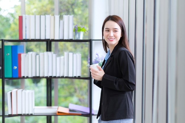 A professional Asian businesswoman is looking out of the window which she holds a coffee cup.