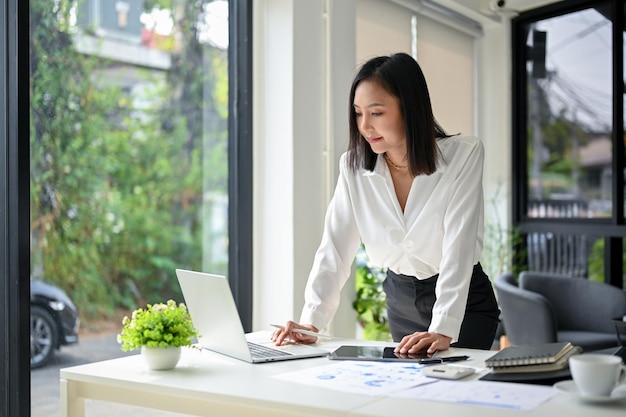 Donna di affari asiatica professionale che si appoggia sul tavolo e utilizza il computer portatile per gestire il suo lavoro