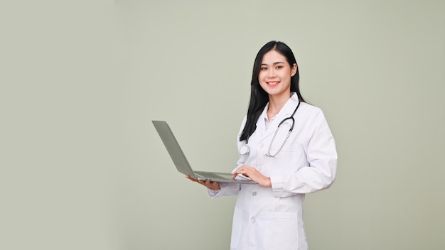 Professional Asian female doctor holding a opened laptop standing against grey background