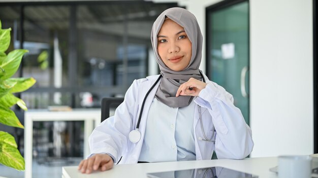 Professional Asian Muslim female doctor in uniform and hijab sits at her desk