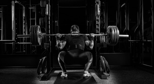 Professional athlete sits with a barbell on his shoulders and prepares to stand with her. View from the back.