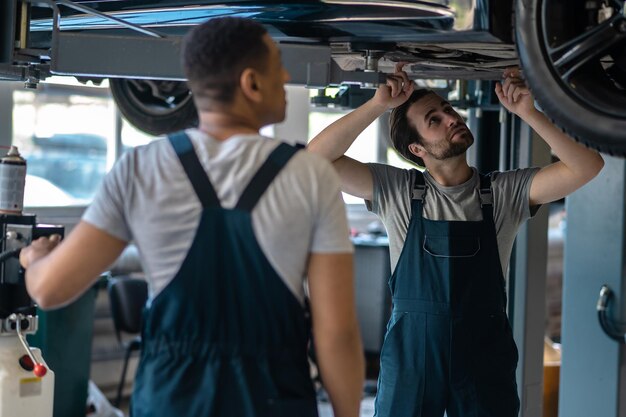 Foto meccanico professionista che ispeziona il sottoscocca sollevato del veicolo sotto la supervisione del suo collega maschio