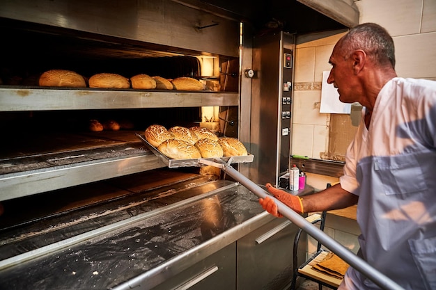 Профессиональный пекарь в униформе достает тележку со свежеиспеченным хлебом из промышленной печи в пекарне