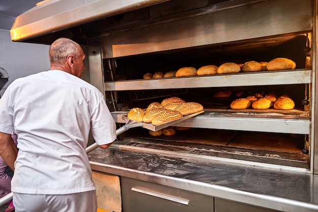 Professional baker in uniform takes out a cart with freshly baked bread from an industrial oven in a bakery