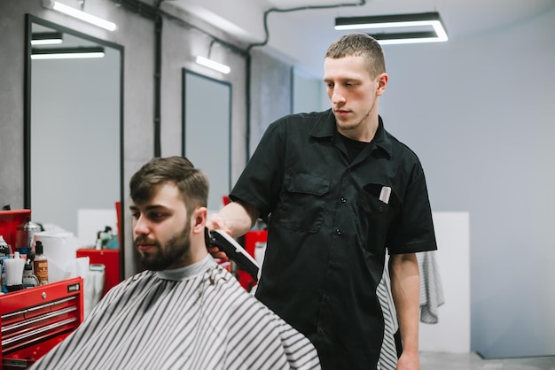 Professional barber cuts the hair of an adult man with a serious face uses a clipper