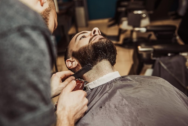 A professional barber cuts his beard to a young hipster man