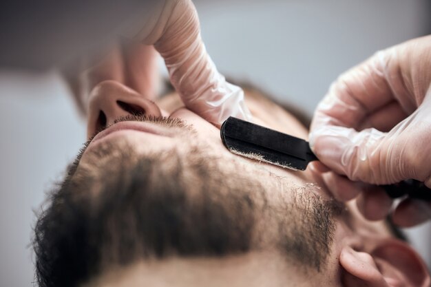 プロの理髪店が理髪店でストレートかみそりを使って若い男にひげを剃る