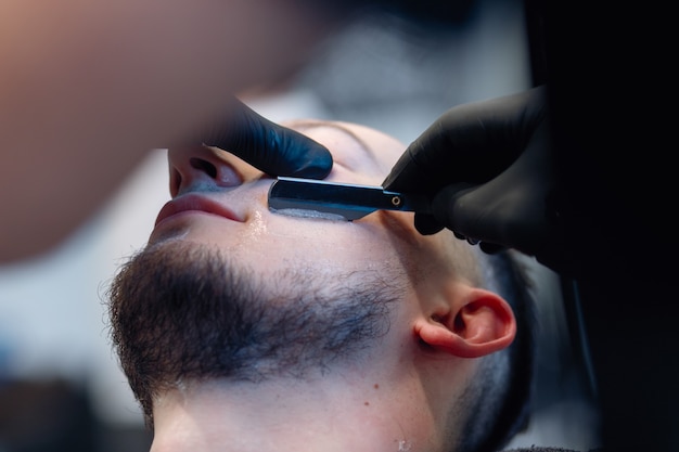 Professional barber shaves customer beard with straight razor. Beard cut with old-fashioned blade at barbershop. Handsome macho man getting his beard shaved in studio. Close-up shot.