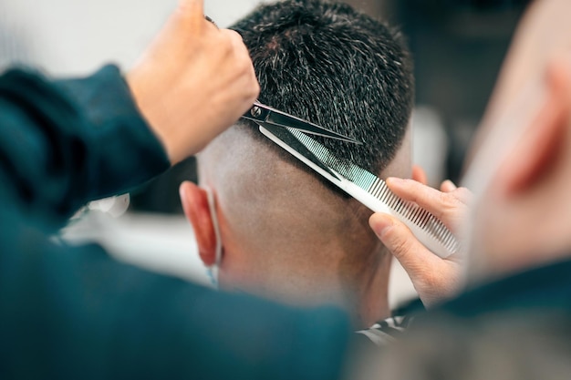 Профессиональный парикмахер, использующий ножницы и расческу, чтобы подстричь мужские волосы в современную модную V-образную прическу, крупным планом вид сзади на его руки и инструменты