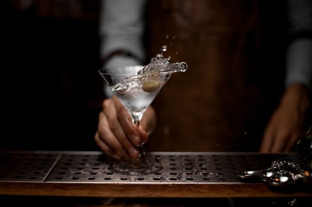 Professional bartender mixing a transparent alcoholic drink in the martini glass with one olive