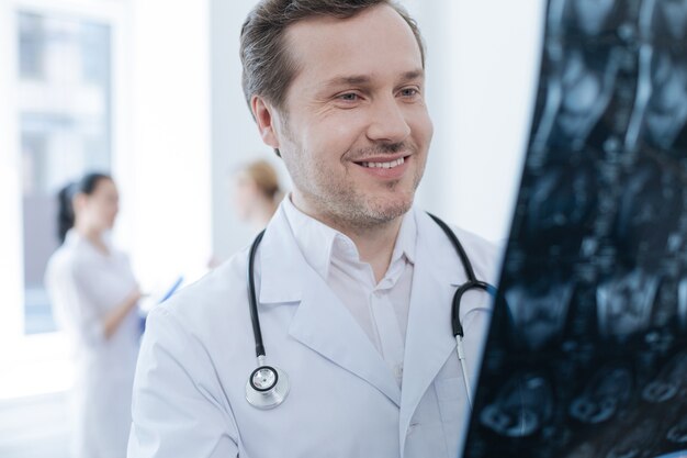 Professional bearded smiling neurosurgeon working at the clinic and holding x ray photo while his colleagues enjoying conversation behind