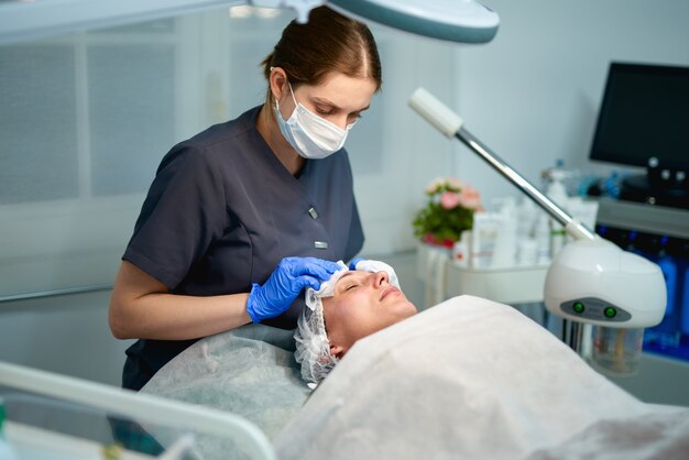 Professional beautician is applying healthy cream on female facial skin