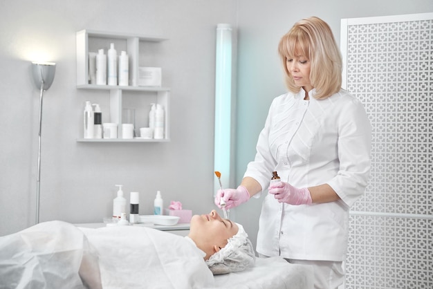 Professional beautician in salon provides comprehensive facial skin care for young woman. Pore cleansing procedures, all-season peeling, firming anti-aging mask