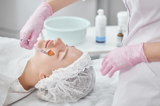 Professional beautician in salon provides comprehensive facial skin care for young woman. Pore cleansing procedures, all-season peeling, firming anti-aging mask