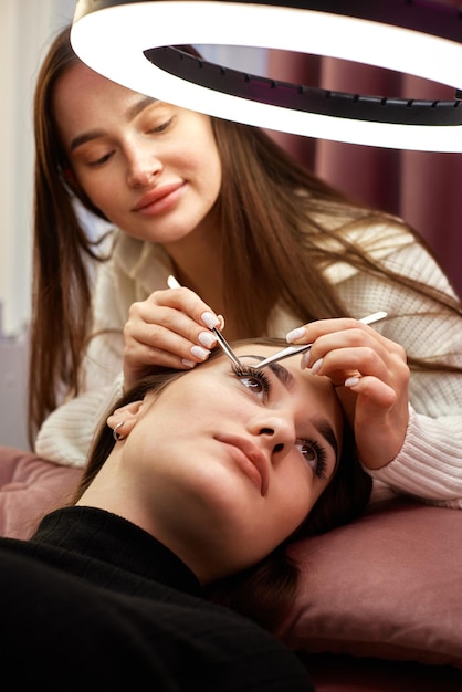 Фото Профессиональный косметолог с пинцетом делает удлинение ресниц для красивой женщины под кольцевым светом
