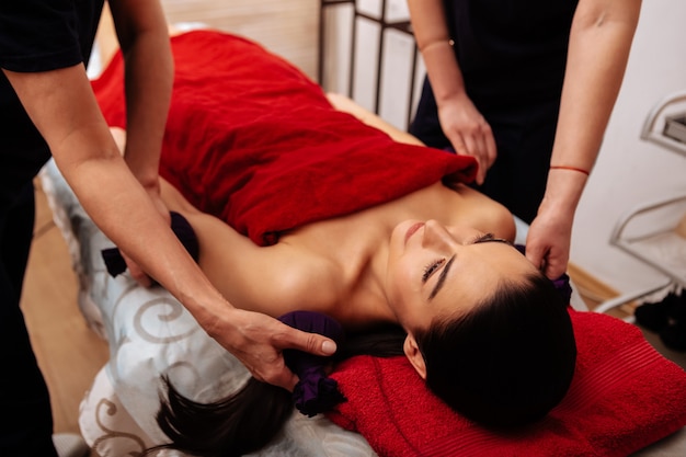 Professional beauty salon. Naked woman covered with red towel resting on massage bed while masters softly stroking her body