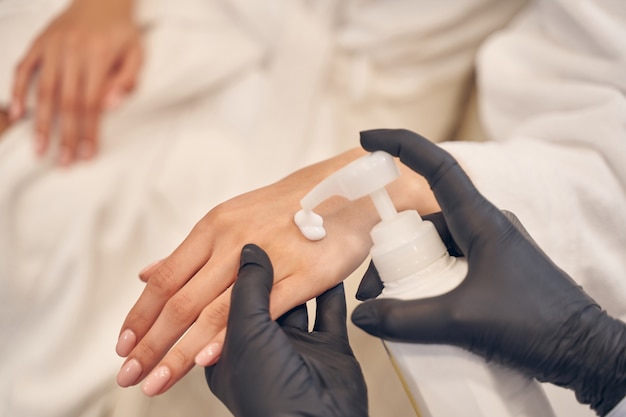 Professional beauty worker applying body lotion on female hand while wearing black gloves