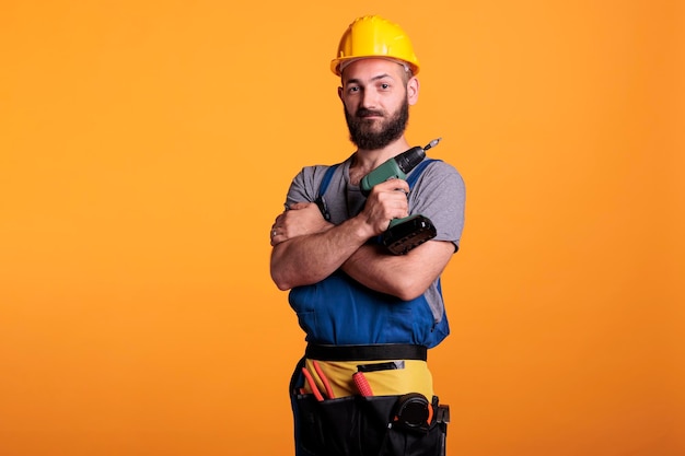 Professional builder holding cordless electric drill, posing in studio. male construction worker using power drilling gun to screw nails, wearing overalls and and hardhat on camera.