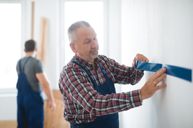 Photo professional builder puts a tape on an empty wall before painting it