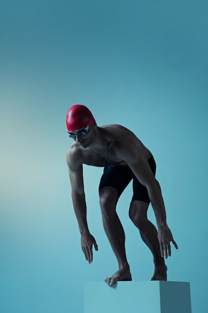 Professional caucasian male swimmer with hat and goggles practicing and training isolated on blue neoned studio background. Grace of motion and action. Healthy lifestyle, sport and movement concept.