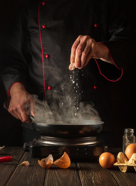 Профессиональный шеф-повар добавляет соль во время приготовления яиц на сковороде Рабочая среда на кухонном столе