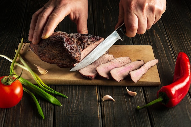 Professional chef cuts the baked beef steak with a knife on a wooden table