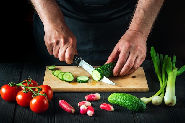 Professional chef cuts a green cucumber on a restaurant kitchen cutting board for salad. Vegetable diet or snack idea.