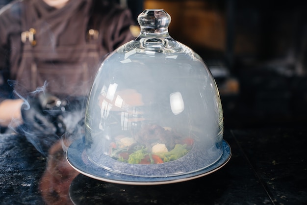 A professional chef serves a freshly prepared salad of tomato and veal greens under a glass hood with thick smoke. Beautiful smoky serving in the restaurant.