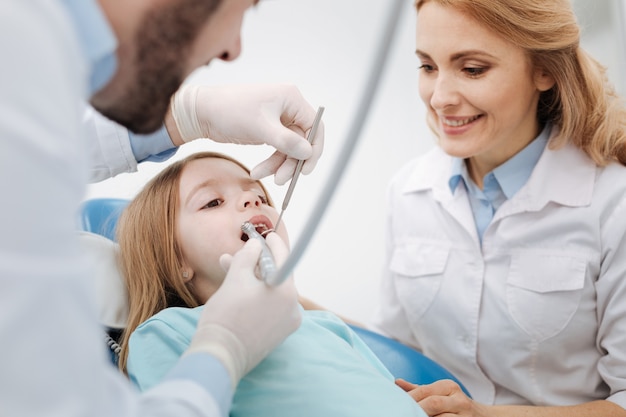 彼の同僚が彼女を慰めている間、小さな患者の歯を調べて定期的な検査を実行している専門の有能な医療の同僚