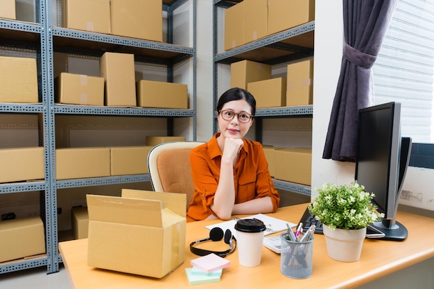 많은 배송 상자가 있는 창고 사무실 책상에 앉아 웃고 있는 카메라를 바라보는 전문적인 여성 온라인 쇼핑 소유자.