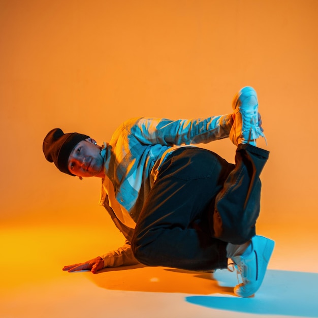 Фото Профессиональный танцор бибой мужчина в модной одежде в шляпе, клетчатой рубашке и кроссовках танцует на полу в творческой цветной студии с оранжевым и синим светом