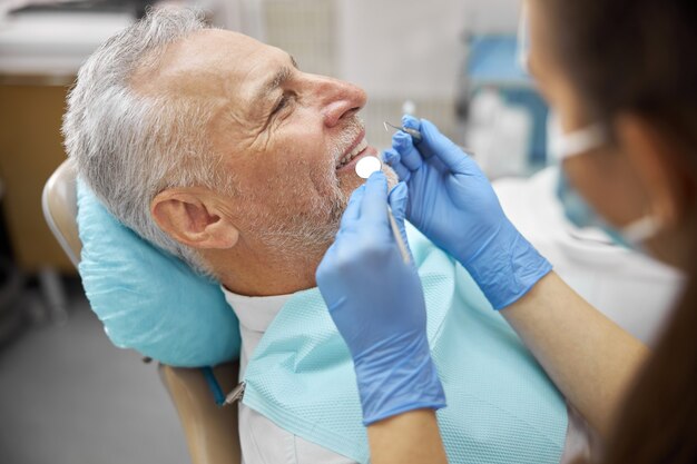 Specialista dentale professionale che tiene uno specchietto per la bocca e un plettro dentale mentre esamina un uomo anziano