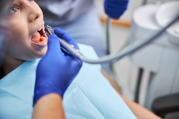 Foto dentista professionista in possesso di un trapano dentale che lo utilizza per il trattamento dei denti di un bambino seduto su una poltrona del dentista