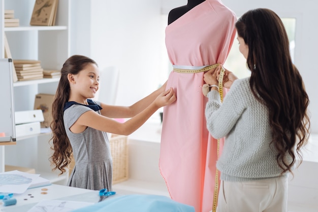 Designer professionista. attraente bella donna intelligente guardando il suo vestito e utilizzando un metro a nastro mentre si lavora insieme a sua figlia