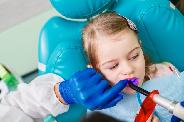 子供の歯科医である専門医は、小さな女の子の歯を器具で治療します。患者診察のための歯科医院。子供の歯科治療のプロセス