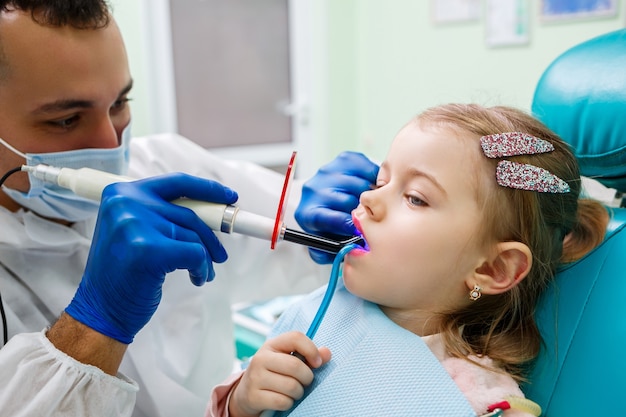 子供の歯科医である専門医は、小さな女の子の歯を器具で治療します。患者診察のための歯科医院。子供の歯科治療のプロセス