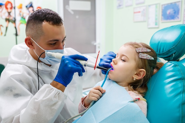 子供の歯科医である専門医は、小さな女の子の歯を器具で治療します。患者診察のための歯科医院。子供の歯科治療のプロセス