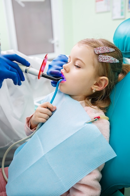 子供の歯科医である専門医は、小さな女の子の歯を器具で治療します。患者診察のための歯科医院。子供の歯科治療のプロセス