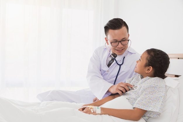 Professional doctor wearing white coat using stethoscope to examine kid patient on bed 